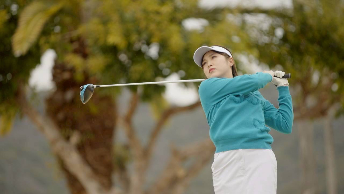 Woman golfer in teal sweater and white cap swings a golf club on a sunny course.