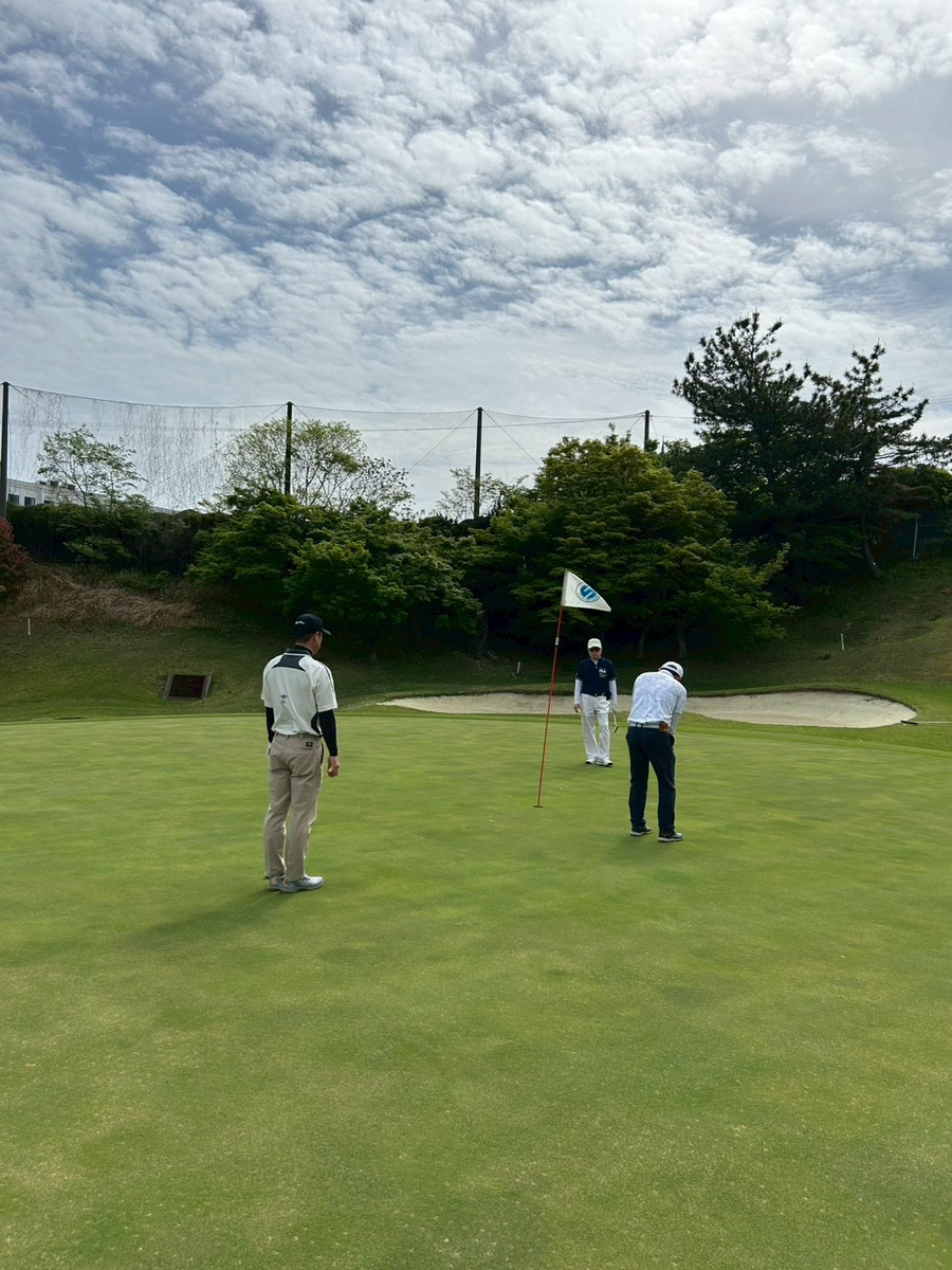 Three golfers on a green putting surface with the flagstick in the hole and a sand bunker in the background under a cloudy sky.