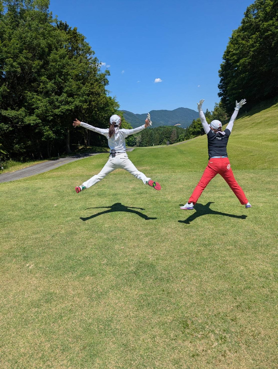 Two people jump mid-air with arms raised on a sunny golf course, green fairways and distant mountains visible in the background.